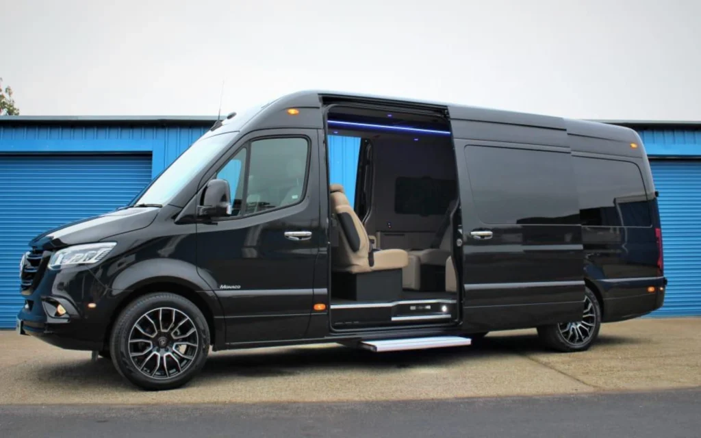 A sleek black Mercedes Jet Class Sprinter van with its side door open, showcasing a luxurious beige interior and ambient lighting.