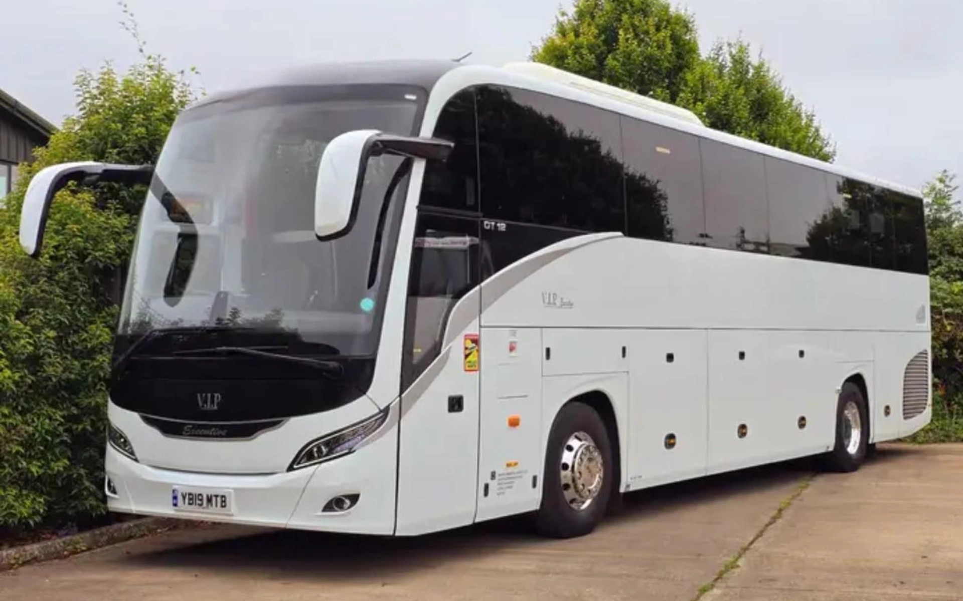 A sleek white luxury coach parked in a driveway surrounded by greenery, showcasing a modern design with tinted windows and a "VIP Executive" label on the front.