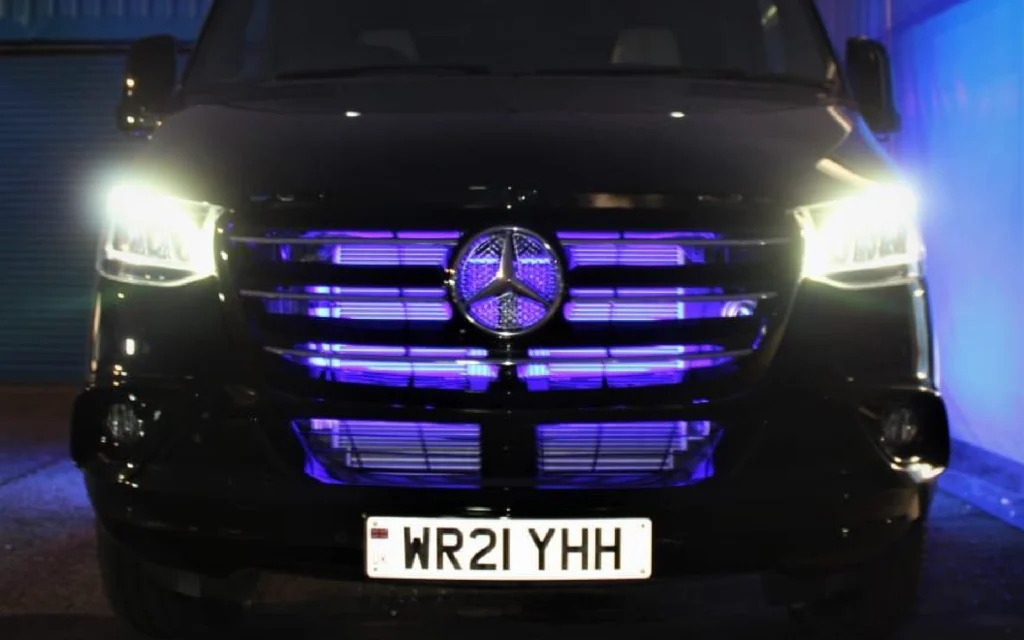 Illuminated front grille of the Mercedes Jet Class Sprinter with glowing blue accents and bright LED headlights, showcasing the vehicle's sleek design and WR21 YHH registration plate.