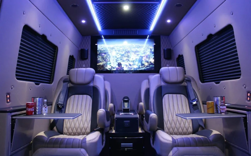 The interior of the Mercedes Jet Class Sprinter with beige quilted leather seats, folding lap tables, ambient lighting, and a large screen displaying a cityscape.