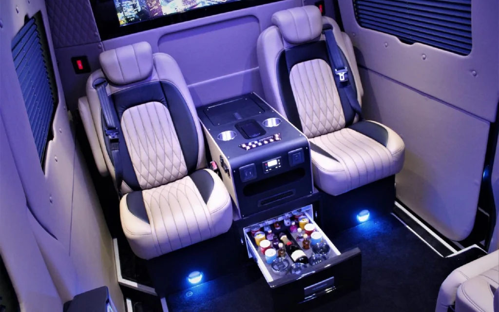 Interior of the Mercedes Jet Class Sprinter featuring two quilted leather seats, a center console with cup holders, and an open beverage drawer stocked with refreshments.