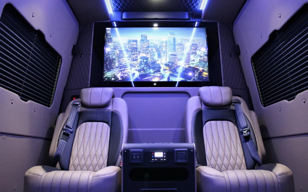 Interior of the Mercedes Jet Class Sprinter featuring two quilted leather seats, a control panel, and a large screen displaying a cityscape.